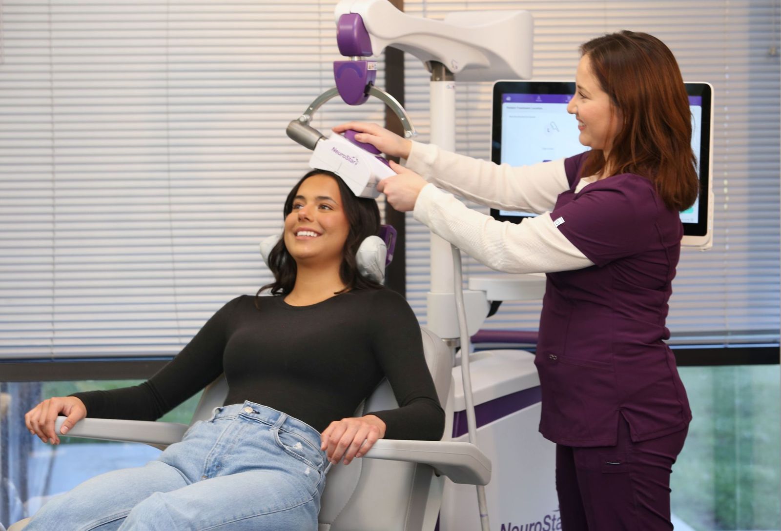 A clinician positions the NeuroStar TMS coil for a smiling patient seated in the treatment chair